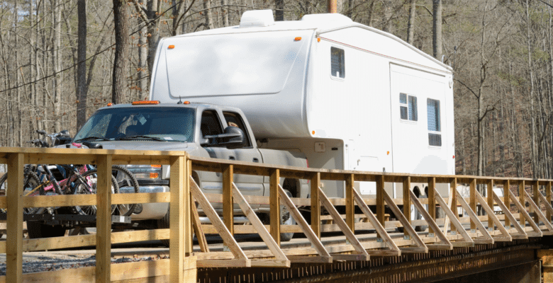 A fifth wheel RV is being towed by a pickup truck across a wooden rail bridge.