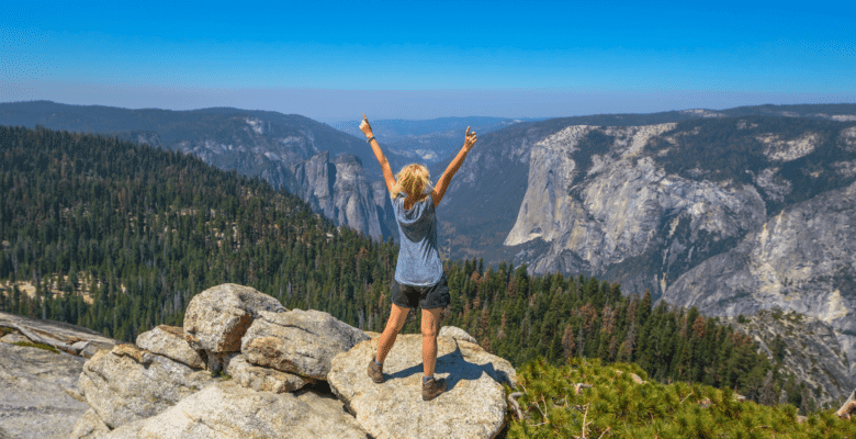 A female hiker stands on a large rock in a valley, surrounded by mountains in Yosemite National Park. She has her hands held high above her head in awe of the view.